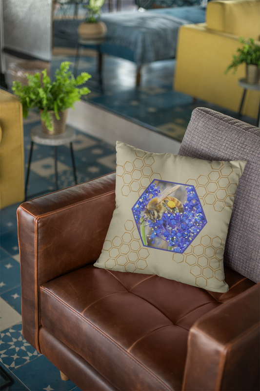Decorative throw pillow with a nature-inspired design featuring a honeybee collecting pollen from vibrant blue Ceanothus flowers, centered in a hexagon motif surrounded by a honeycomb pattern. Displayed on a brown leather armchair in a modern living room with stylish decor.